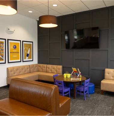 Children play area in dental office with leather sofas, purple kid-sized chairs around a small activity table, framed artwork on the wall, and a mounted TV